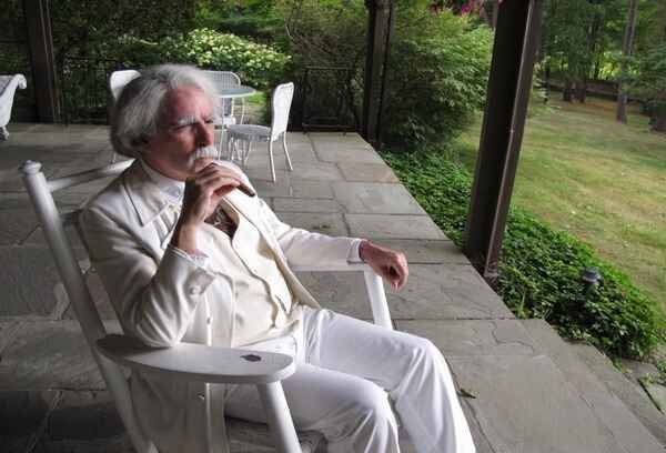 Mark Dawidziak in a white suit dressed as Mark Twain sitting in a rocking chair on an outside porch.