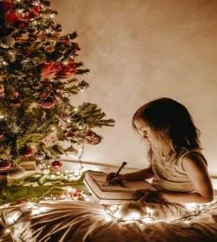 Little girl sitting beside a lighted Christmas tree while drawing on paper.
